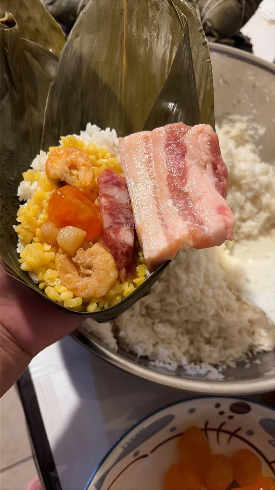 An open bamboo-leaf zongzi being filled by hand in You Yi’s kitchen — rice and mung beans layered with dried shrimp, salted egg yolk, sausage, and a slab of pork belly.